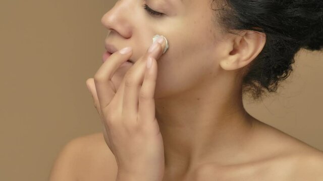 Beauty portrait of young African American woman applying skincare clay mask on her face. Black female model posing on brown studio background. Close up. Slow motion ready, 4K at 59.94fps.