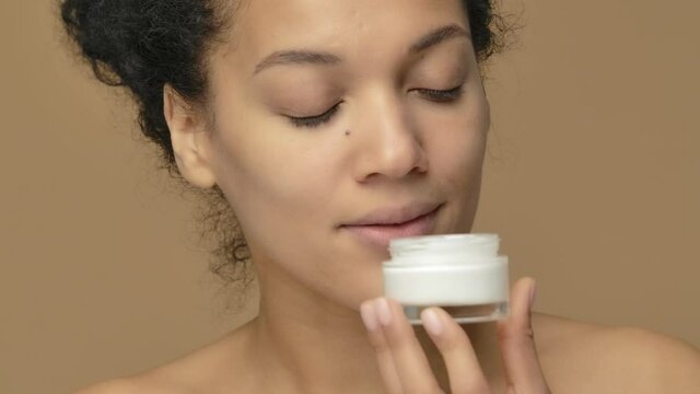 Beauty portrait of young African American woman twisting off a jar of cream and enjoying its aroma. Black female model posing on brown studio background. Close up. Slow motion ready, 4K at 59.94fps.