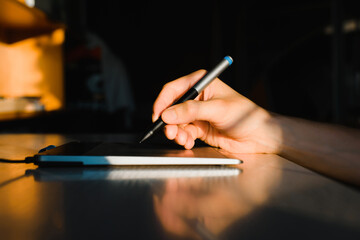 Indoor designer working on a professional tablet with a pen, at sunrise or sunset. Close-up of a man's hand holding a graphic pen for drawing