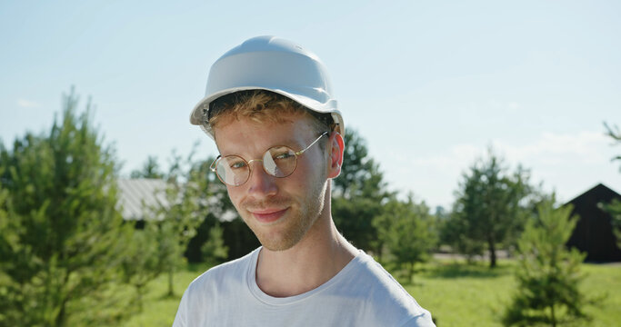 Cheerful Young Man In Glasses Taking Off Hardhat And Touching Hair While Resting During Work In Yard On Summer Day