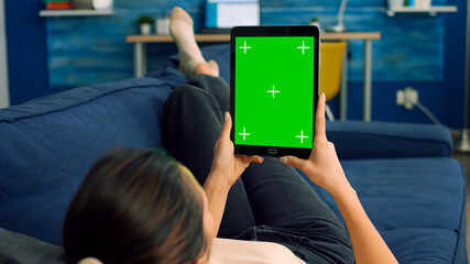Casual woman lying on sofa using tablet computer mock up green screen chroma key in vertical portrait mode. Freelancer browsing on internet with isolated device for online project