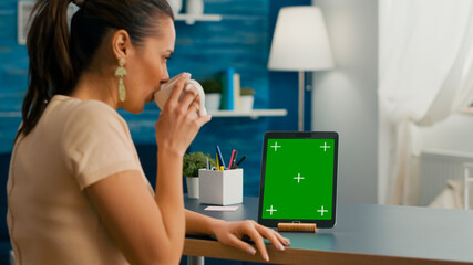 Freelancer woman holding cup of coffee looking at tablet computer with mock up green screen chroma key sitting on desk table. Caucasian female browsing on isolated device from home office room