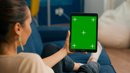 Caucasian female lying on sofa having online meeting on tablet computer with mock up green screen chroma key display. Woman using isolated touchscreen device for social networks browsing