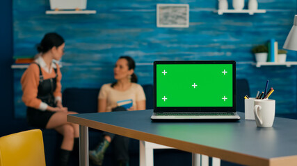 Laptop computer with mock up green screen chroma key standing on desk while in background two women...