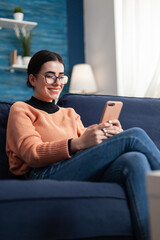 Smiling student relaxing on couch while talking with her friends on message about social media communication. Teenager woman chatting on social network using mobile app device - obrazy, fototapety, plakaty