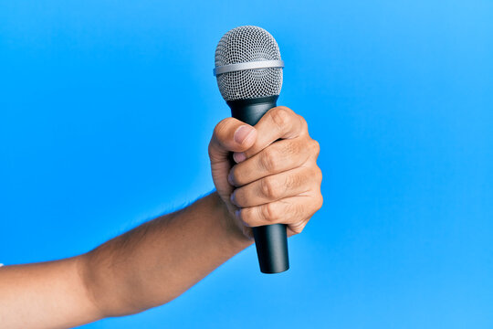 Hand Of Hispanic Man Holding Microphone Over Isolated Blue Background.