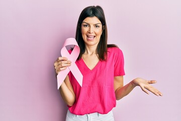Young hispanic woman holding pink cancer ribbon celebrating achievement with happy smile and winner...