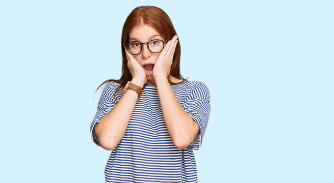 Young read head woman wearing casual clothes and glasses afraid and shocked, surprise and amazed expression with hands on face
