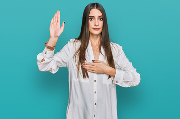 Young beautiful woman wearing casual white shirt swearing with hand on chest and open palm, making...
