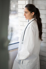 Side view of the dreaming young doctor who is standing indoor and looking through the window