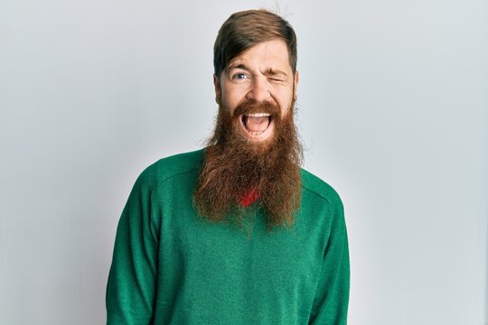 Redhead man with long beard wearing casual clothes winking looking at the camera with sexy expression, cheerful and happy face.