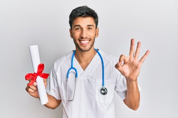 Young handsome man wearing doctor uniform holding medical degree doing ok sign with fingers,...