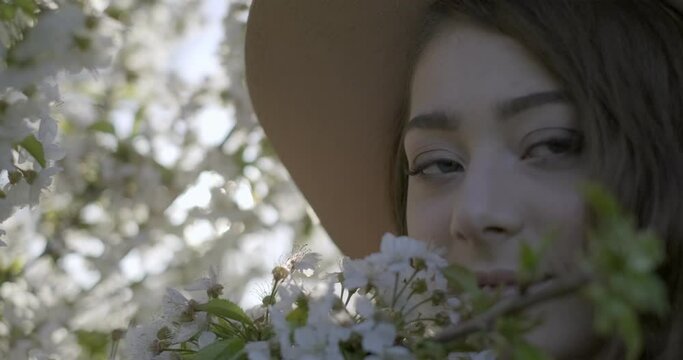 Close-up of brown hared girl with grey eyes wearing a hat and smelling the flowering branch of apple tree. Filmed outdoors. Blurred background