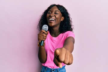 Beautiful african young woman singing song using microphone pointing to camera smiling and laughing hard out loud because funny crazy joke.