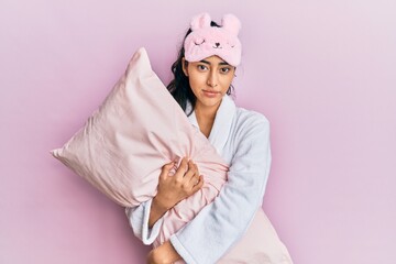 Hispanic teenager girl with dental braces wearing sleep mask and robe hugging pillow relaxed with serious expression on face. simple and natural looking at the camera.