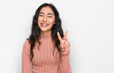 Hispanic teenager girl with dental braces wearing casual clothes showing and pointing up with fingers number two while smiling confident and happy.