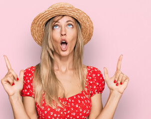Beautiful caucasian woman wearing summer hat amazed and surprised looking up and pointing with...