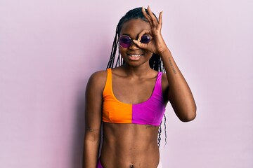 Young african american woman wearing bikini and sunglasses doing ok gesture with hand smiling, eye looking through fingers with happy face.