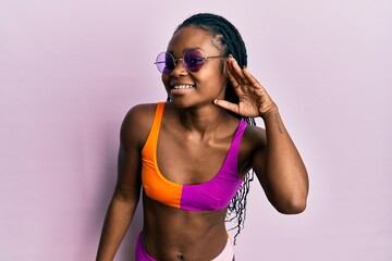 Young african american woman wearing bikini and sunglasses smiling with hand over ear listening an hearing to rumor or gossip. deafness concept.