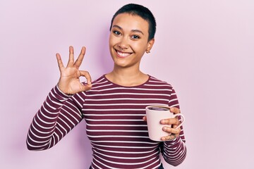 Beautiful hispanic woman with short hair drinking a cup of coffee doing ok sign with fingers, smiling friendly gesturing excellent symbol