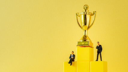 Miniature of businessman stands on podium ladder with golden trophy on yellow background