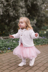 Cute blonde toddler girl in a pink skirt and a white sweater. Running outdoors. Background of spring park.