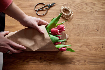 Female hands arranging pink tulips bouquet on wooden table, floristic hobby workplace