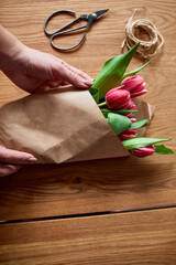 Female hands arranging pink tulips bouquet on wooden table, floristic hobby workplace