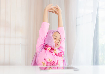 Obraz premium Cheerful Muslim girl relaxing at comfortable office chair stretching arms in the air, Asian kid girls study online via on a tablet while in the living room at the home, kids and education concepts