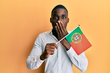 Young african american man holding portugal flag covering mouth with hand, shocked and afraid for...