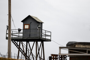An old gray observation tower made of sheet metal with lantern spotlights and a window shot in the military industrial zone in Russia against the white cloudy sky of pillars and a concrete fence.