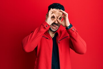 Young hispanic man wearing red leather jacket doing ok gesture like binoculars sticking tongue out, eyes looking through fingers. crazy expression.