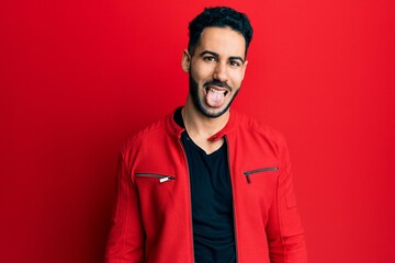 Young hispanic man wearing red leather jacket sticking tongue out happy with funny expression. emotion concept.
