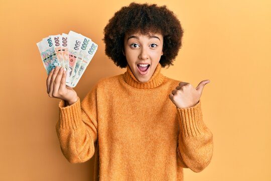 Young hispanic girl holding czech koruna banknotes pointing thumb up to the side smiling happy with open mouth