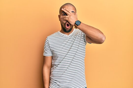 Hispanic adult man wearing casual clothes peeking in shock covering face and eyes with hand, looking through fingers afraid