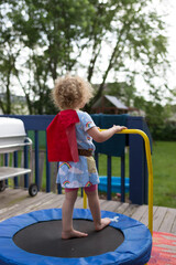 Fototapeta na wymiar Little girl superhero on a trampoline in the backyard; diy costume on blonde curly haired child