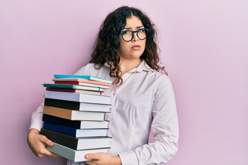 Young brunette woman with curly hair holding a pile of books in shock face, looking skeptical and sarcastic, surprised with open mouth