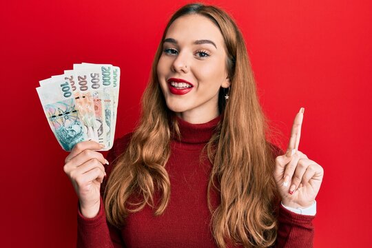 Young blonde woman holding czech koruna banknotes smiling with an idea or question pointing finger with happy face, number one