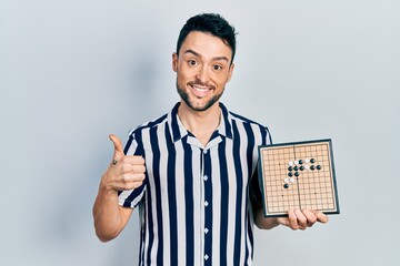 Young hispanic man holding asian go game board smiling happy and positive, thumb up doing excellent and approval sign