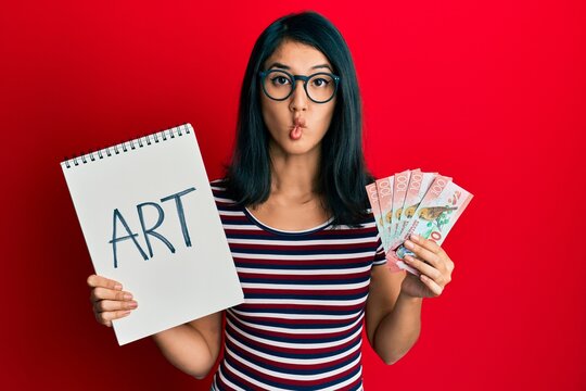Beautiful asian young woman holding art notebook and 100 new zealand dollars making fish face with mouth and squinting eyes, crazy and comical.