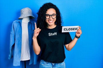 Young hispanic woman with curly hair wearing staff t shirt holding closed banner looking at the camera smiling with open arms for hug. cheerful expression embracing happiness.