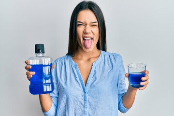 Young hispanic girl holding mouthwash for fresh breath sticking tongue out happy with funny...