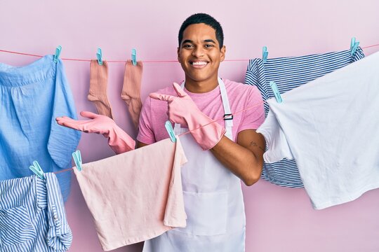 Young handsome hispanic man wearing cleaner apron holding clothes on clothesline amazed and smiling to the camera while presenting with hand and pointing with finger.