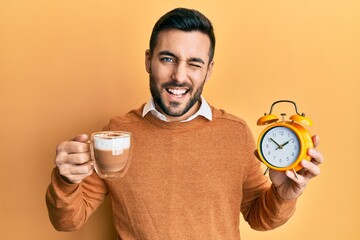 Young hispanic man holding coffee and alarm clock winking looking at the camera with sexy...