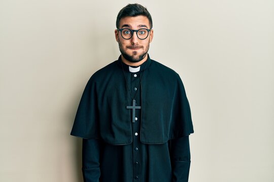 Young hispanic man wearing priest uniform standing over white background puffing cheeks with funny face. mouth inflated with air, crazy expression.