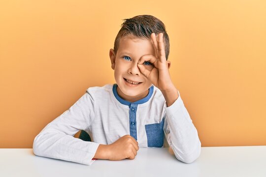 Adorable caucasian kid wearing casual clothes sitting on the table doing ok gesture with hand smiling, eye looking through fingers with happy face.