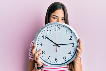 Young brunette woman holding big clock covering face smiling with a happy and cool smile on face....