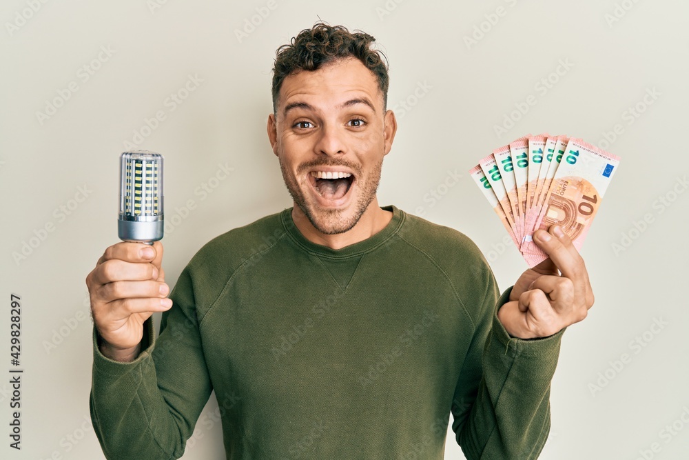 Sticker Young hispanic man holding led bulb and euros banknotes celebrating crazy and amazed for success with open eyes screaming excited.