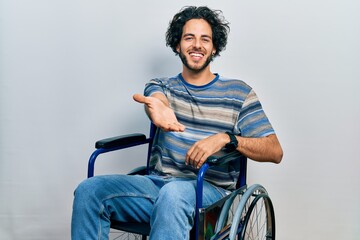Handsome hispanic man sitting on wheelchair smiling cheerful offering palm hand giving assistance...