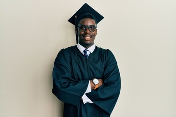 Handsome black man wearing graduation cap and ceremony robe happy face smiling with crossed arms...